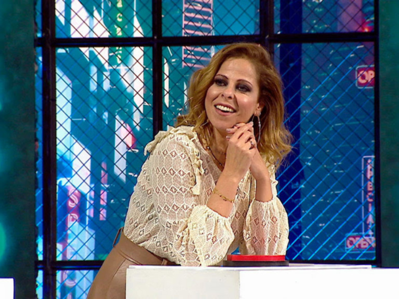 A una mujer sonriendo mientras se sienta en un escritorio. El fondo es una pantalla con letras y símbolos de colores brillantes, probablemente en un estudio de televisión.