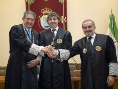 Tres hombres en trajes formales se saludan con una sonrisa, frente a un fondo de un escudo y una bandera.