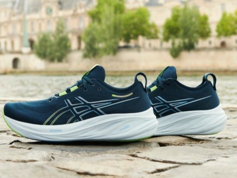 Zapatillas de running azul marino con detalles en verde y blanco, sobre un suelo de piedra. Fondo borroso con árboles y edificios en el horizonte.