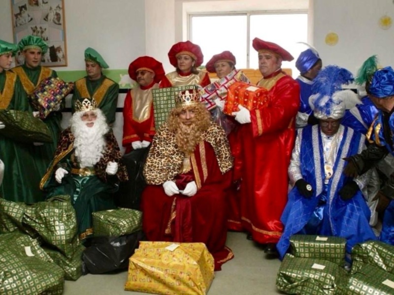 Los Reyes Magos de Oriente, vestidos con trajes tradicionales y sombreros rojos, son sorprendidos por un grupo de niños que les entregan regalos. La escena se desarrolla en una habitación con un lienzo de Navidad y una ventana que permite la entrada de luz natural. Los regalos variados en colores, como verde y amarillo, están dispuestos sobre el suelo.