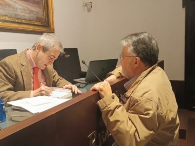 Dos hombres sentados en una sala de oficinas, uno escribiendo y el otro observando. La imagen muestra un ambiente profesional con elementos como una computadora, documentos y una silla de oficina.