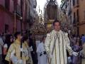 Una procesión religiosa con personas vestidas de blanco y dorado, una imagen de la Virgen en el centro, y edificios típicos de la ciudad al fondo.