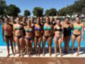 Equipo femenino de natación posando frente a una piscina azul, vestidos con trajes de baño coloridos.