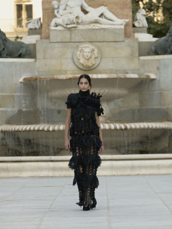 Una mujer con un vestido negro de encaje camina frente a una fuente histórica. La imagen muestra un paisaje urbano con elementos antiguos y modernos, destacando la moda y el arte.