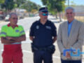 Tres personas, dos de ellas con uniforme de policía y una en traje formal, posan frente a una calle con árboles y un edificio al fondo. La imagen parece ser de un evento o reunión oficial en una ciudad.