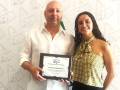 Un hombre y una mujer son felices mostrando un premio de la IFAAM. El hombre sostiene el premio mientras sonríe, y la mujer está a su lado con una sonrisa amigable. El fondo es blanco con patrones gráficos de IFAAM.