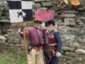 Dos hombres vestidos con trajes medievales posan junto a una bandera y un cartel en un entorno histórico.