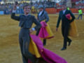 Un torero con una capa colorida y un chaleco amarillo se prepara para la matanza de toros en una plaza de toros. El público observa desde las gradas y un toro está visible en el fondo.