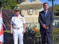 Dos hombres, uno en uniforme naval y otro en traje formal, posan frente a un monumento con columnas. El paisaje incluye árboles, palmeras y un cielo azul claro.