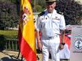 Un hombre en uniforme militar español, posando con una bandera española y una bandera de la Armada en un entorno urbano.