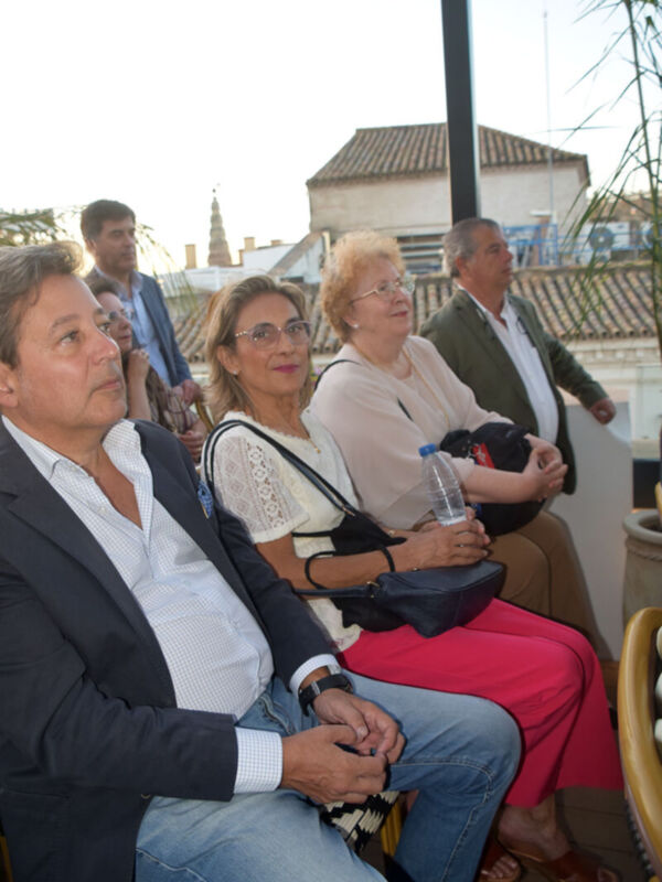 Tres personas sentadas en un evento, con una mujer en el centro y dos mujeres a sus lados. La imagen muestra detalles de la vestimenta y una mano con un bolso negro. En el fondo, se puede ver una estructura arquitectónica y un cielo despejado.