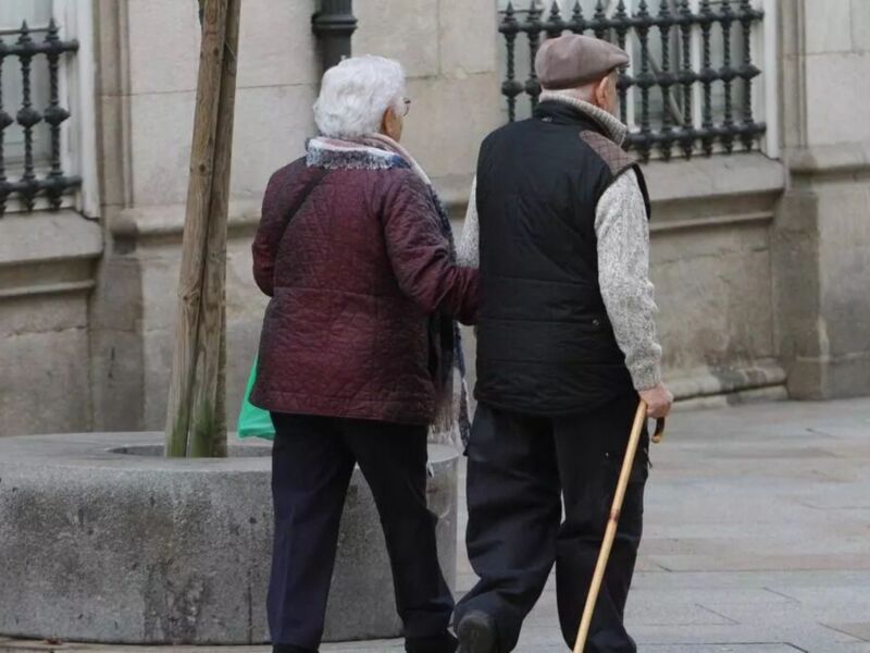 Dos personas caminan juntas, una con abrigo rojo y la otra con chaqueta negra. Detrás de ellos, una columna verde se alza en un entorno urbano con una barandilla metálica.