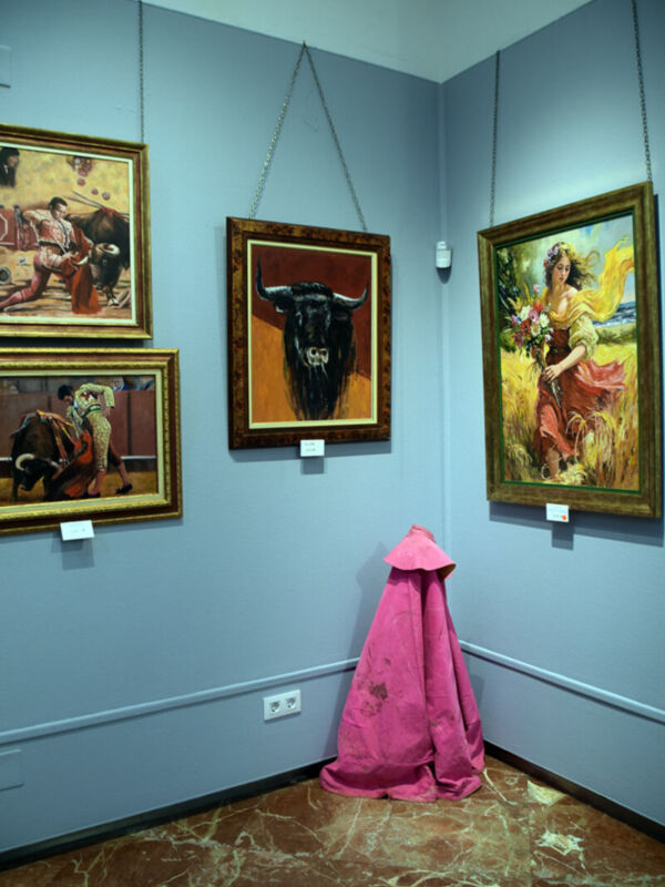 Una galería de arte con tres cuadros enmarcados colgando en la pared. El primero es un retrato de una figura histórica vestida con trajes tradicionales, el segundo es una pintura de un toro en un fondo rojo, y el tercero muestra a una mujer con vestido largo y flores en un paisaje. Debajo de los cuadros, se observa una cortina roja y un interruptor eléctrico en la pared. El suelo es de mármol con patrones geométricos.