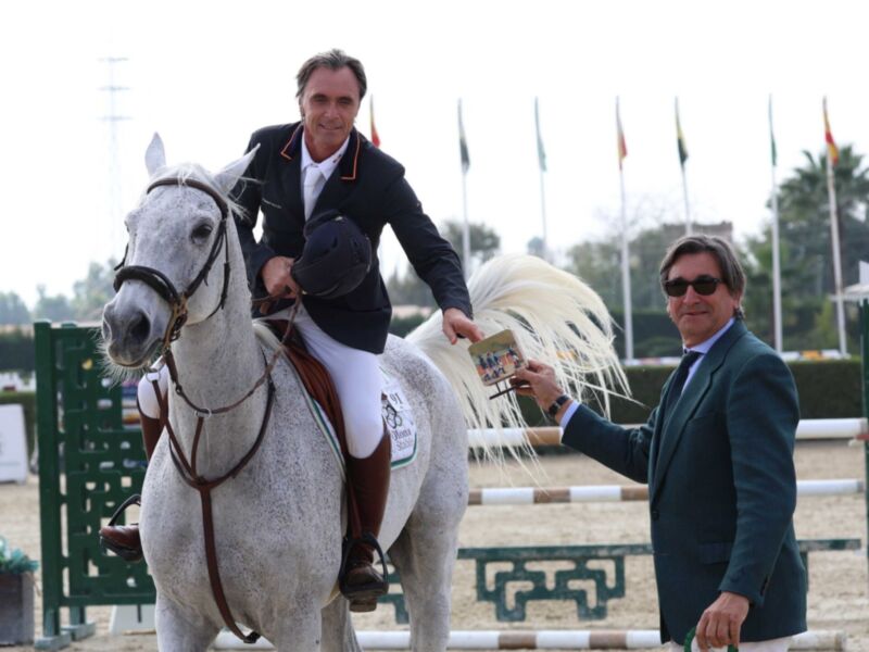 Un hombre monta un caballo blanco en una pista de salto, con otro hombre a su lado sosteniendo un trofeo. El caballo tiene una cinta blanca en la cola y el hombre monta está usando un traje negro con detalles rojos. En el fondo, hay banderas y palmeras en un ambiente de competición ecuestre.