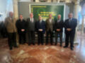 A seis hombres vestidos formalmente, posando frente a un cartel que anuncia eventos celebrativos por los 20 años de existencia y más de 100 años de historia. La fecha mencionada es del 24 al 30 de noviembre de 2025.