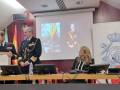 A tres personas en uniforme militar, posiblemente oficiales o militares, celebrando una ceremonia. En el fondo se puede ver un proyector con una imagen de un oficial militar y la bandera nacional española. La escena parece ser parte de una conferencia o evento oficial, con un fondo que incluye elementos institucionales.