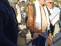 Un grupo de personas caminando por una calle con edificios y árboles en el fondo. Algunos llevan bufandas y chaquetas, mientras que otra persona sostiene una botella grande. La escena parece ser en un día soleado y la gente está caminando en diferentes direcciones.