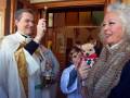 Una imagen festiva donde un sacerdote celebra una bendición mientras una mujer sostiene a un pequeño perro. La escena está ambientada en una iglesia con detalles decorativos y personas de diferentes edades participando activamente.